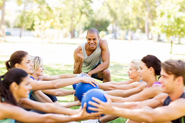 Personal Trainer Motivating People Exercising With Medicine Ball Young male personal trainer motivating people exercising in bootcamp workout class with medicine balls in park. Horizontal shot. military camp stock pictures, royalty-free photos & images