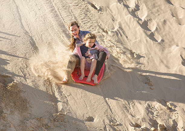 A teenager sandboards down a sand dune in summer.