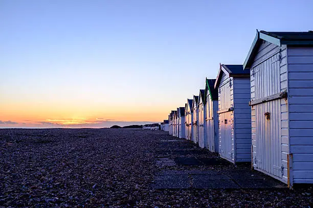 landscape image of a row of white beach huts on the right of the frame leading off into the distance with diminishing perspective  with multi coloured roof trims on a pebbled beach at sunset