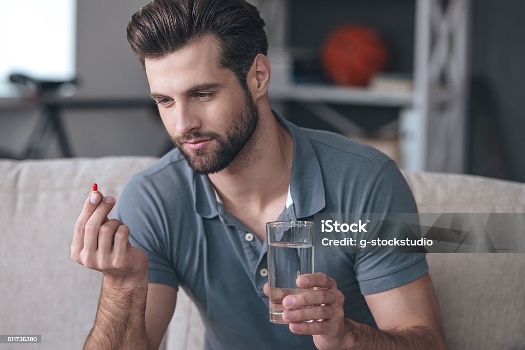 Just one pill can help. Handsome young man holding a glass of water and looking at a pill in his hand while sitting on the couch at home Men Stock Photo