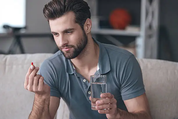 Handsome young man holding a glass of water and looking at a pill in his hand while sitting on the couch at home