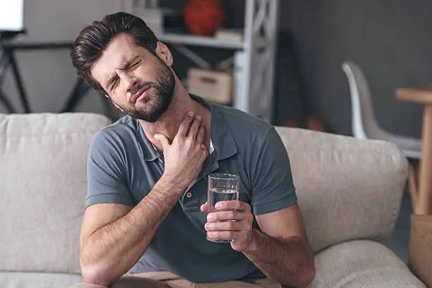 Frustrated handsome young man touching his neck and holding a glass of water while sitting on the couch at home