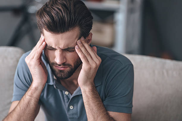 Feeling stressed. Frustrated handsome young man touching his head and keeping eyes closed while sitting on the couch at home headache photos stock pictures, royalty-free photos & images
