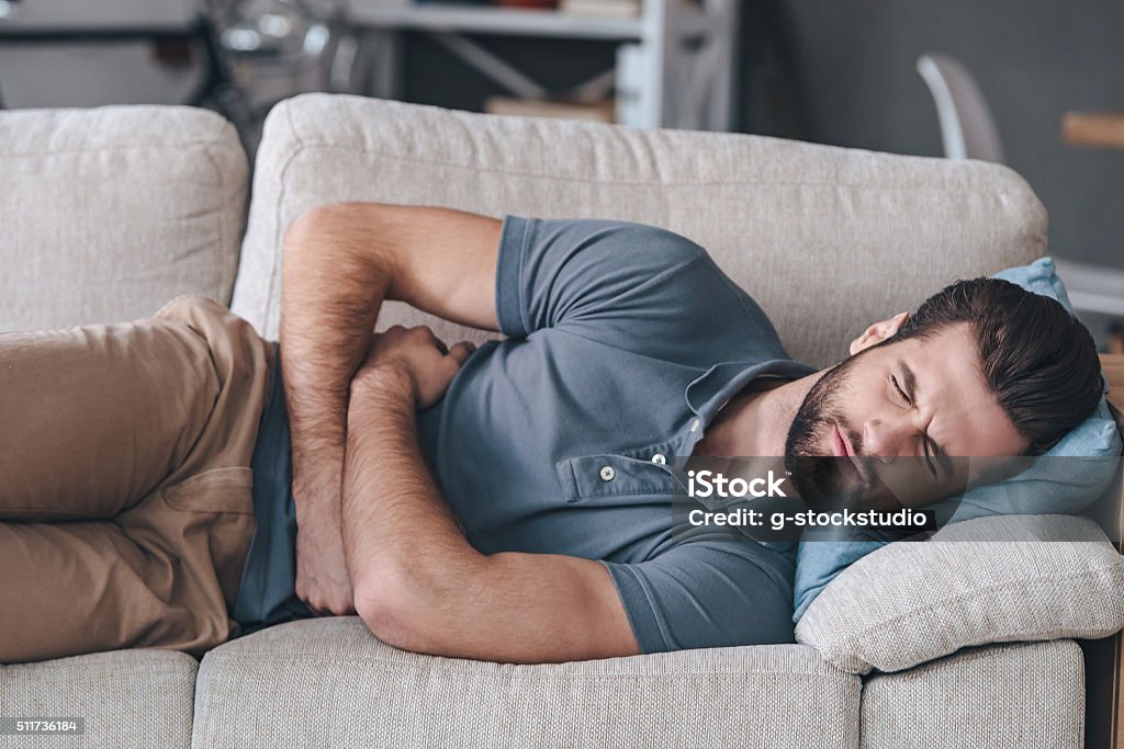 Terrible stomachache. Frustrated handsome young man hugging his belly and keeping eyes closed while lying on the couch at home Stomachache Stock Photo
