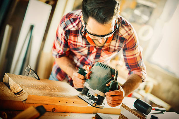 Carpentry workshop. Closeup front view of handsome early 30's man doing carpentry work in his garage. His scraping edges of a plank with a electric plunge router . Wearing protective glasses and lumberjack clothes. Tilt shot. plane hand tool stock pictures, royalty-free photos & images