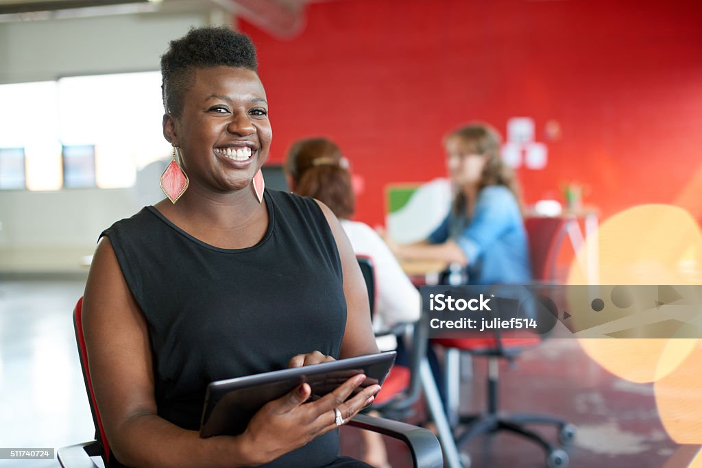 Confident female designer working on a digital tablet in red Casual portrait of an african american business woman using technology in a bright and sunny startup with the team in the background African Ethnicity Stock Photo