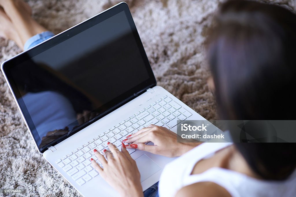 Hands of a woman typing on laptop computer High angle over the shoulder view of the hands of a woman typing on a laptop computer as she sits on the carpet at home 20-24 Years Stock Photo