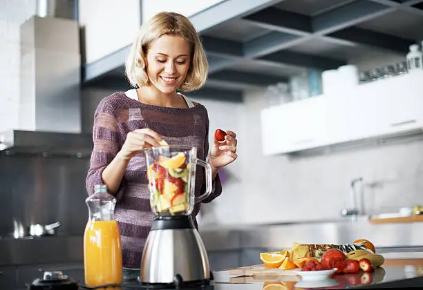 A young woman smiling happily while placing slices of fresh fruit into her blender