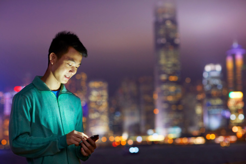Shot of a young man holding a cellphone in front of the Hong Kong skyline at night