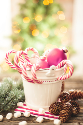 Candy canes in a small white pail and other christmas decorations on a wooden table. Christmas tree on the background. 