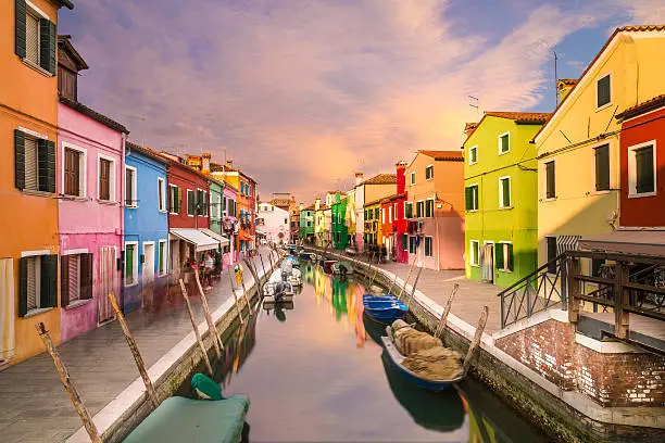 A view of colourful houses in Burano during the day along the canal. Colourful clouds can be seen in the sky.