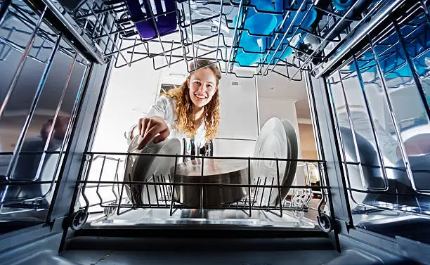 A smiling young woman seen, unusually, from inside the dishwasher drum, loads or unloads dishes.