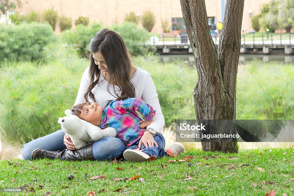 Mother and daughter Young colombian mother and her daughter looking at each other and sitting on the grass on a city park. Latin Script Stock Photo