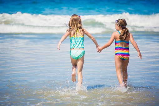 Two cute little girls enjoying an endless summer at the beach. Running hand in hand to the water together view from behind