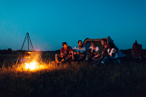 Group of young hikers camping. They enjoy camping and having fun. They are sitting around campfire, smiling and talking. Fist pot is on fire on front and tent is behind them in background. Deliblatska pescara, Bela Crkva, Serbia, Europe