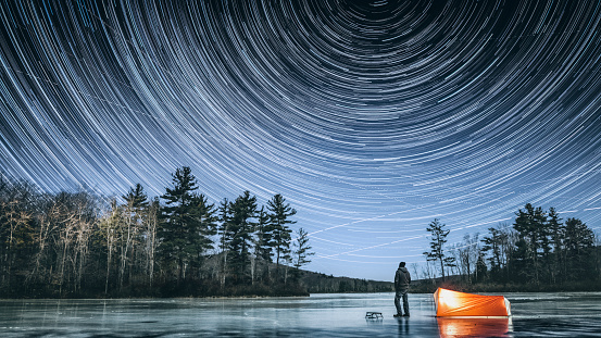 Frozen in time man standing on the icy lake in Connecticut's dark and wild north west corner of the state, next to his tent looking up observing star trails on cold full moon winter night.