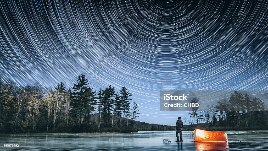 Winter stargazing in Connecticut Frozen in time man standing on the icy lake in Connecticut's dark and wild north west corner of the state, next to his tent looking up observing star trails on cold full moon winter night. Winter Stock Photo