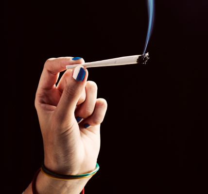 A woman's elegantly manicured hand holds up a smoking hand-rolled spliff or joint of marijuana.