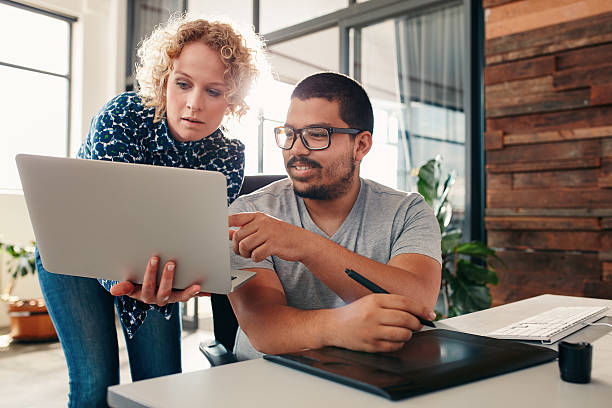 Graphic designers cooperating in design studio Portrait of young man sitting at his desk pointing at laptop in hands of a female colleague, male graphic designer help a female coworker in office. young graphic designer stock pictures, royalty-free photos & images