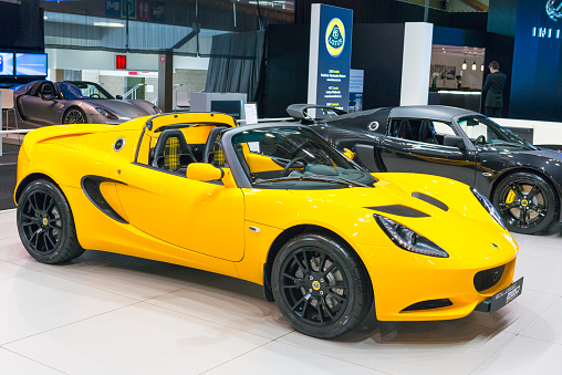 Brussels, Belgium - Januari 12, 2016: Yellow Lotus Elise Sport 220 sports car front view. The Elise Sport is a super lightweight version of the Lotus Elise sports car. The car is on display during the 2016 Brussels Motor Show. The car is displayed on a motor show stand, with lights reflecting off of the body. There are people looking around and other cars on display in the background.