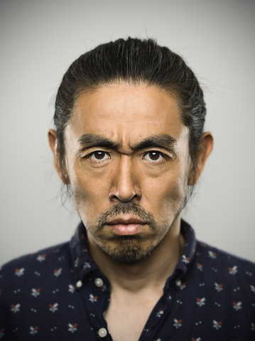 Studio portrait of a japanese man looking at camera with negative expression. The man has around 40 years and has long hair and casual clothes. Vertical color image from a medium format digital camera. Sharp focus on eyes.