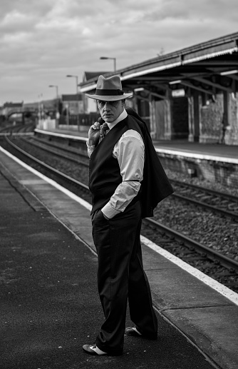 Black and white vertical image of a mature man dressed as a 1940s gangster charcater standing by train tracks.