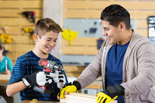 Preteen uses power drill in local woodworking shop. Father or instructor at woodworking day camp holds boards steady as boy prepares to use the drill. The boy is confidently smiling. Both are wearing protective gloves.