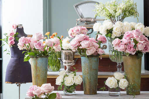 A scene from a flower shop.  You can see containers and vases of flowers on a tiered display.  The flowers are almost all white and pink.  Most of the flowers are roses.  There is nobody in the photo.
