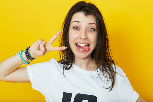 Cheerful lovely girl showing her tongue and peace gesture over yellow background