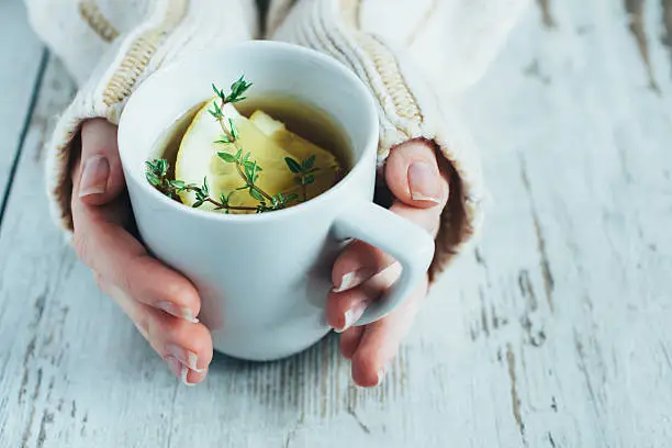 Human hands holding cup of tea with thyme herb and lemon slices on a wooden table,.