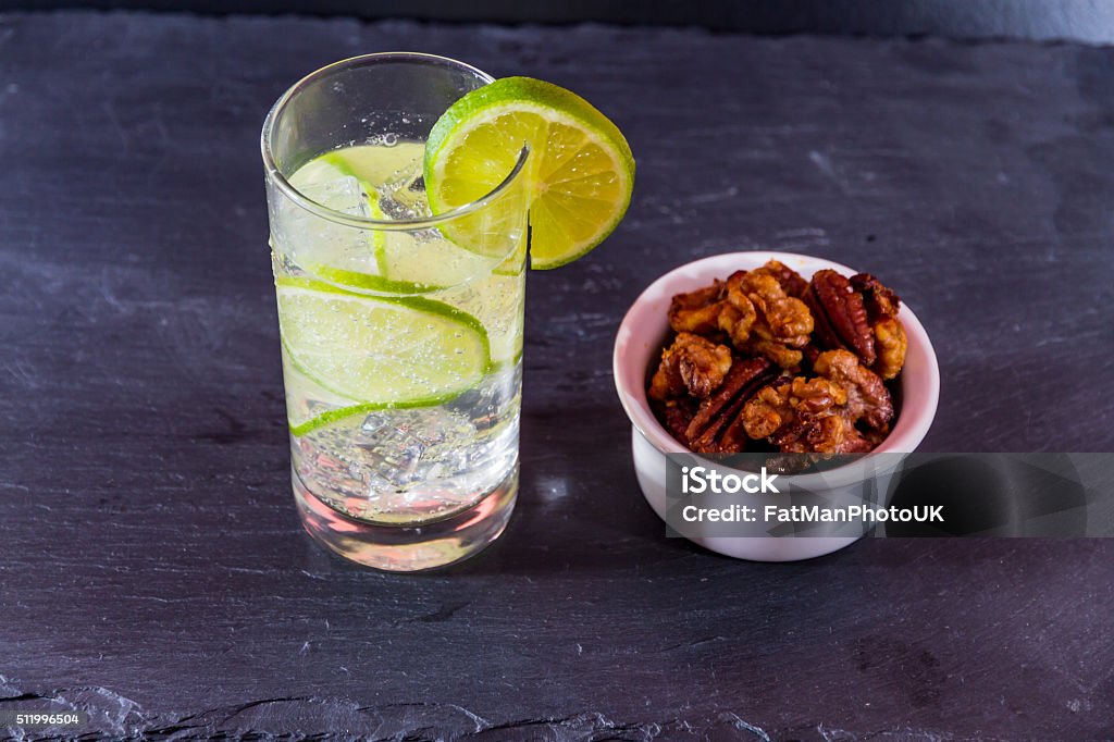 Gin and tonic with bowl of mixed roast nuts. Gin and tonic garnished with lime, served with bowl of home roasted nuts. Alcohol - Drink Stock Photo