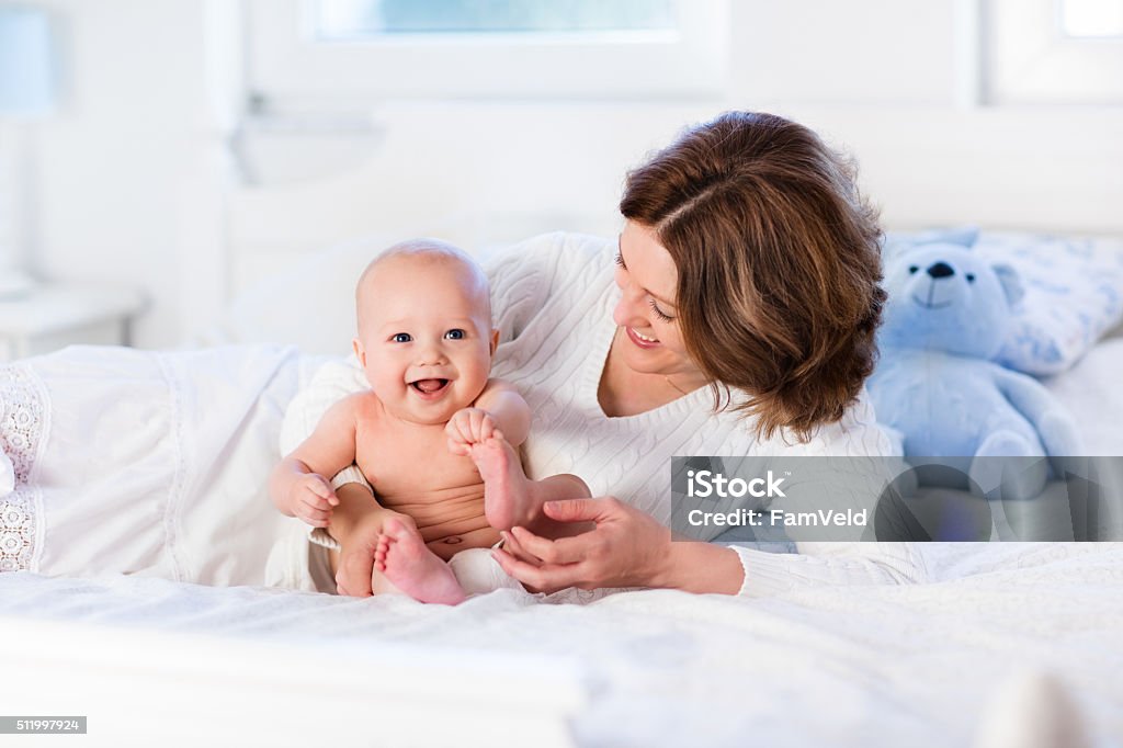 Adorable mother and baby on a white bed Mother and child on a white bed. Mom and baby boy in diaper playing in sunny bedroom. Parent and little kid relaxing at home. Family having fun together. Bedding and textile for infant nursery. Baby - Human Age Stock Photo