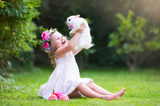 Girl playing with real rabbit in sunny garden. Child and bunny on Easter egg hunt in flower meadow. Toddler kid feeding pet animal. Kids and pets play.  Fun and friendship for animals and children.
