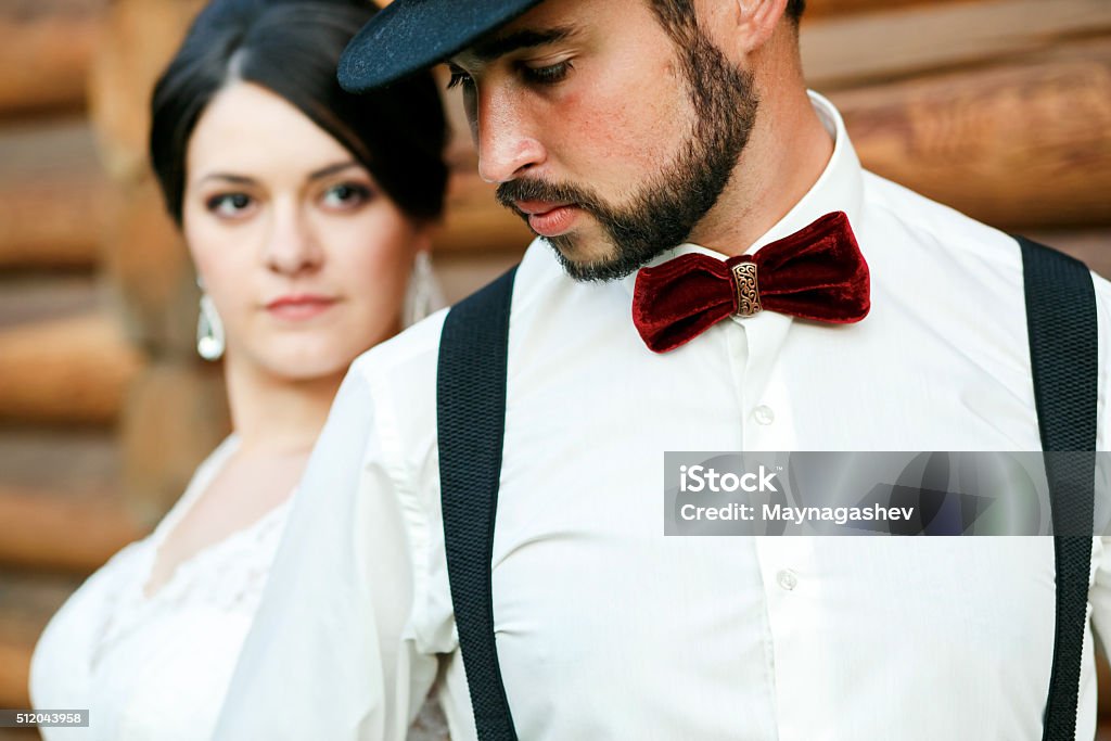 Thoughtful groom in hat with beard, mustache, bow tie and Thoughtful groom in hat with beard, mustache, bow tie and suspenders. Bride in white wedding dress. Stylish couple standing near wall of timber, looking away, close-up portrait. Gangster style. Adult Stock Photo