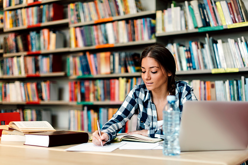 Female Student sitting in library searching information on laptop for her assignment. She is sitting at table surfing internet for notes for her study. Writing in her notepad.