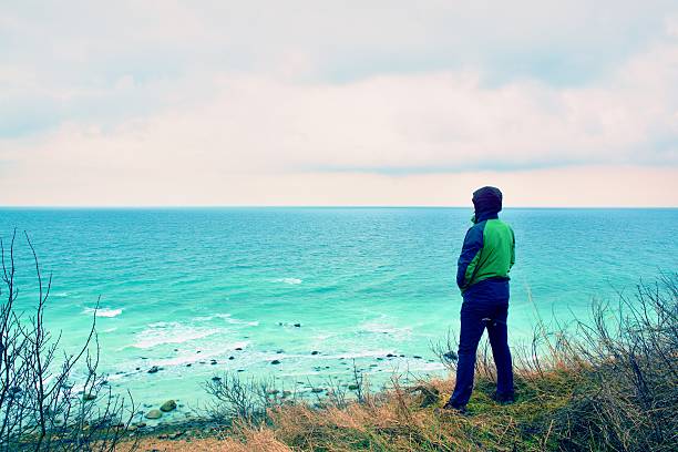 istock Man in green windcheater on cliff look at sea horizon. 512057762