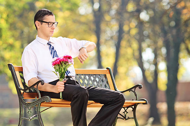Young man holding flowers and checking the time seated on bench in park