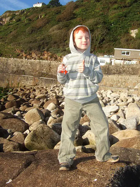 Photo showing a young boy on a beach with large pebbles / rocks.  The boy is pictured wearing a grey hoodie (hood up), with his thumbs up as he poses for the camera whilst eating a bar of chocolate.