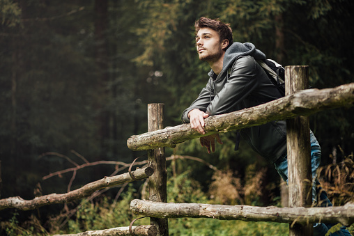 Young man with backpack hiking in the forest and leaning on a wooden fence, nature and physical exercise concept