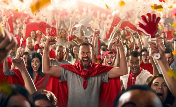 On the foreground a group of cheering fans watch a sport championship on stadium. A man in the centre stands with his hands up to the sky. Everybody are happy. People are dressed in casual cloth. Colourful confetti flies int the air.