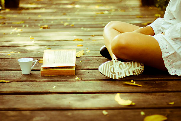 Portrait of a beautiful elegant young woman sitting outdoors barefoot and reading a book in the park or garden on a bright sunny day of summer on the green grass background
