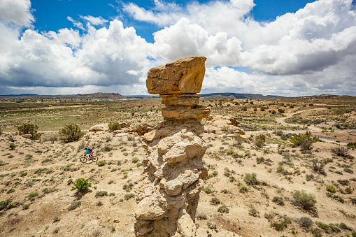 a woman pedals past a rock formation underneath a cloud filled blue sky.  horizontal wide angle composition taken in gallup, new mexico.