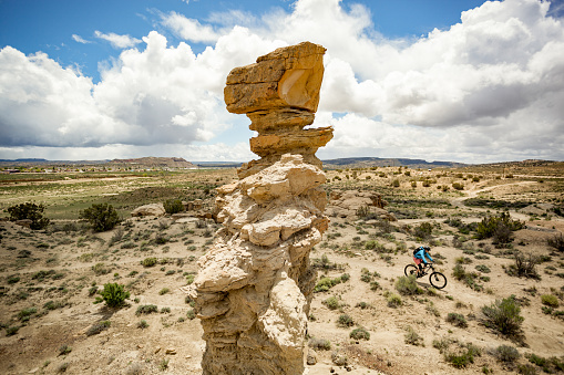 a woman pedals past a rock formation underneath a cloud filled blue sky.  horizontal wide angle composition taken in gallup, new mexico.