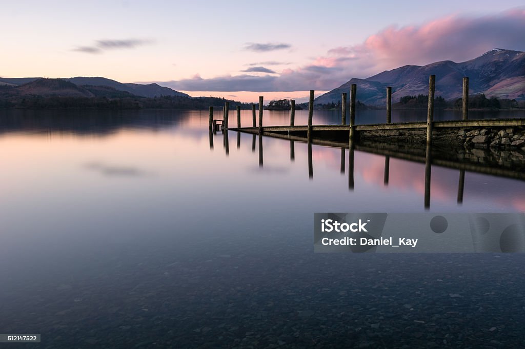 Jetty Reflections In Calm Lake With Pink Clouds In Sky. Ashness Jetty reflecting in a calm Derwentwater lake in Keswick, Lake District, UK. Taken on a cold winter evening the photograph shows a simple but nice winter dusk sunset with a subtle pink/purple colour in the clouds. Zen-like Stock Photo