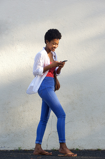 Full length side portrait of a happy young african woman walking on street and reading text message on mobile phone