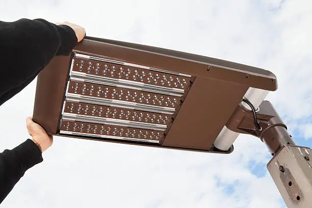 A worker, standing in an elevated platform, is installing an LED street light, adjusting the final position against an overcast sky. This could work equally well for a parking lot light concept.