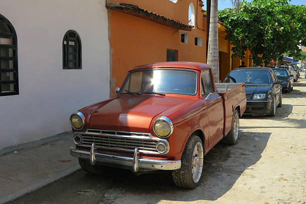 Shot of the front of an old red brown datsun truck parked on a dusty road, in front of a row of cars and palm trees, outside an old Spanish hacienda. Complete with vintage front bumper, grille and headlights. Taken in Puerto Vallarta, Mexico.
