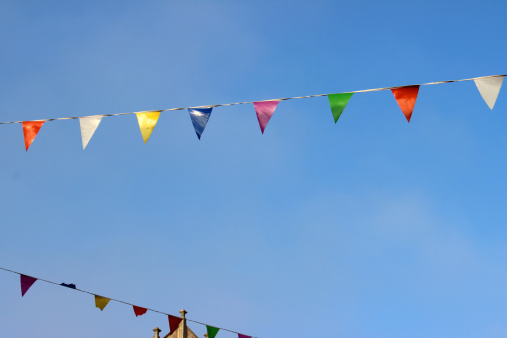 Photo showing a row of rainbow coloured bunting flags on a tight wire rope, against a blue sky.  The flags have been put up as part of local celebrations - as the carnival is coming to town.