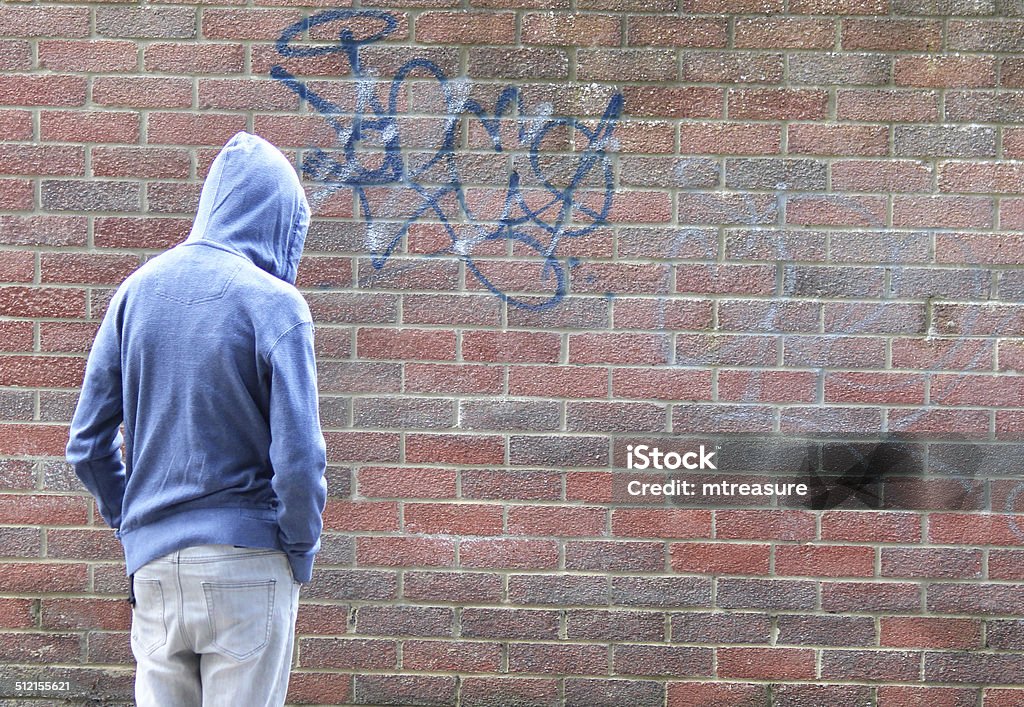 Image of teenage boy / youth wearing hoodie, beside graffiti brick-wall Photo showing a teenage boy / youth wearing a blue hoodie and standing beside a dirty brick wall with graffiti, in a run-down part of the city. Hooded Shirt Stock Photo