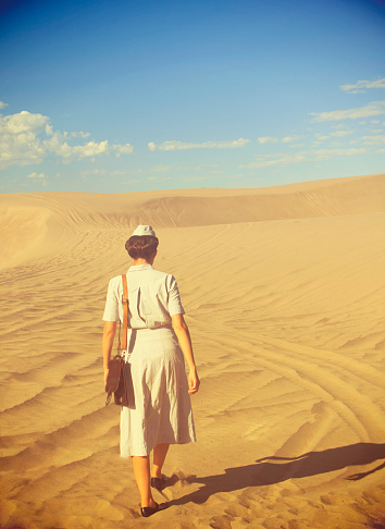 Young WWII Nurse Walking In The Desert With Her back To The Camera. She wears an authentic female medical dress uniform and carries a leather shoulder bag. There is lots of copy space in the sky